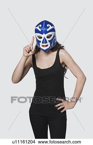 Picture - Young female wearing wrestling mask as she raises index finger against gray background. Fotosearch