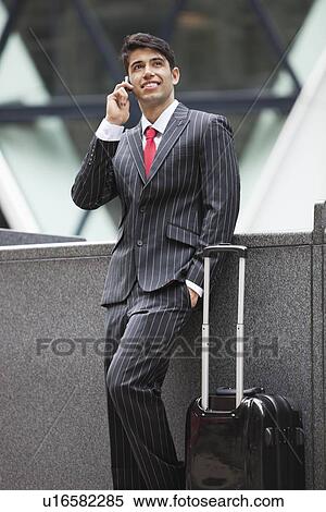 Young Indian businessman communicating on cell phone while standing next to luggage bag View Large Photo Image Stock Photography - Young Indian businessman communicating on cell phone while standing next to luggage bag. Fotosearch