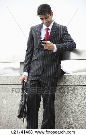 Young Indian businessman texting through cell phone while holding laptop bag at parapet View Large Photo Image Stock Photo - Young Indian businessman texting through cell phone while holding laptop bag at parapet. Fotosearch