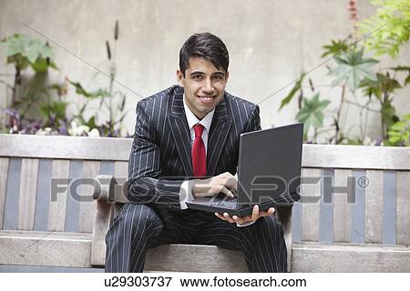 Young Indian businessman using laptop while sitting on bench View Large Photo Image Stock Photo - Young Indian businessman using laptop while sitting on bench. Fotosearch