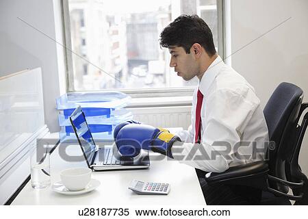 Stock Photography - Young Indian businessman wearing boxing gloves while using laptop at office desk. Fotosearch