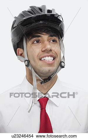 Picture - Young Indian businessman wearing cycling helmet while looking away against gray background. Fotosearch