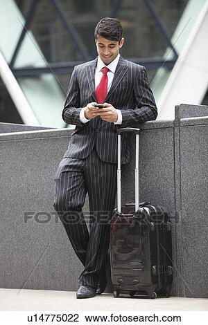 Stock Image - Young Indian businessman with luggage bag using cell phone. Fotosearch