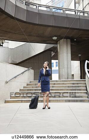 Stock Photograph - Young Indian businesswoman using cell phone with luggage outdoors. Fotosearch