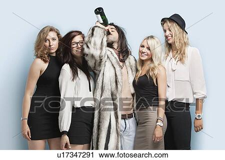 Stock Image - Young man drinking beer while standing with female friends against light blue background. Fotosearch