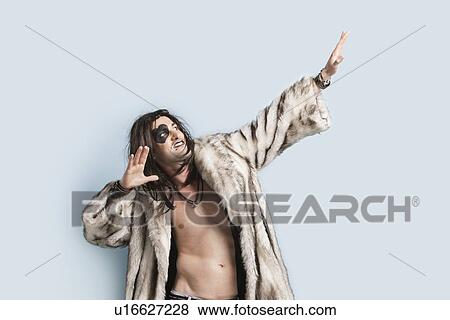 Stock Photo - Young man in fur coat looking up with arms raised against light blue background. Fotosearch