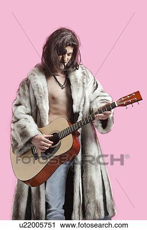 Stock Image - Young man in fur coat playing guitar against pink background. Fotosearch