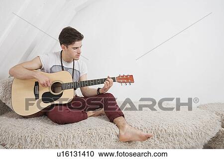 Stock Image - Young man playing guitar while sitting on fur sofa. Fotosearch