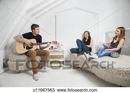 Stock Image - Young man playing guitar with female friends sitting on sofa. Fotosearch