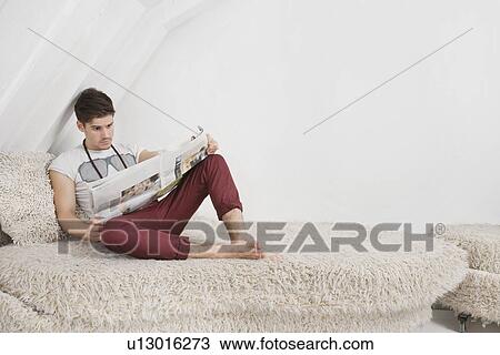 Stock Image - Young man reading newspaper while sitting on fur sofa. Fotosearch