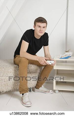Stock Photo - Young man sitting on fur ottoman with video game controller. Fotosearch