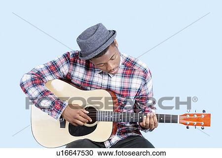 Stock Image - Young man wearing hat as he plays guitar over light blue background. Fotosearch