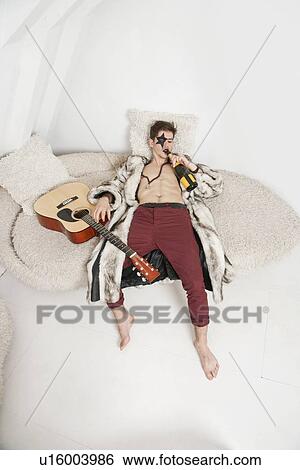 Stock Photograph - Young man with guitar drinking while lying on sofa. Fotosearch