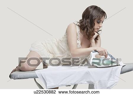 Stock Image - Young woman comfortably ironing shirt against gray background. Fotosearch