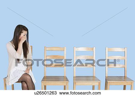 Stock Image - Young woman crying besides empty wooden chairs over blue background. Fotosearch