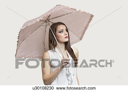 Young woman holding umbrella while looking away over gray background View Large Photo Image Stock Image - Young woman holding umbrella while looking away over gray background. Fotosearch