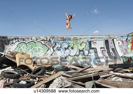 Stock Photo - Young woman in bikini jumping over graffiti wall with garbage in foreground. Fotosearch