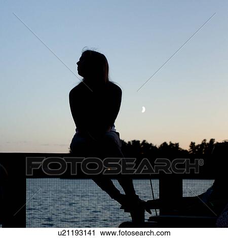 Stock Image - Silhouette of a woman sitting on a railing, Lake of the Woods, Ontario, Canada. Fotosearch