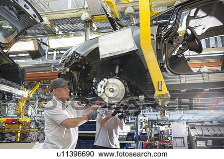 Stock Image - Car assembly workers fitting axles to cars on production line in car factory. Fotosearch