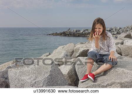 Stock Image - Girl eating apple on rocky beach. Fotosearch
