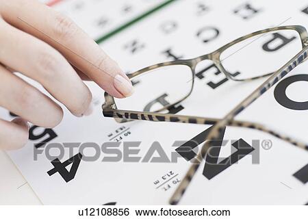 Stock Photograph - Close up of woman's hands holding glasses above eye chart, studio shot. Fotosearch