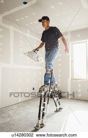 Stock Image - Drywall worker on stilts walking across unfinished room. Fotosearch