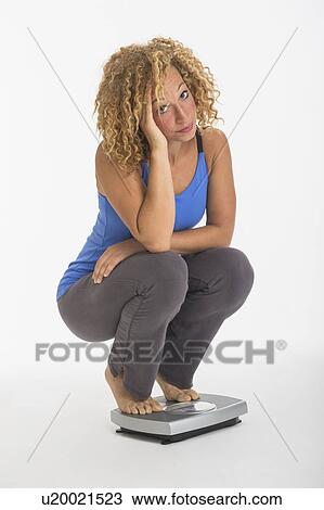 Stock Image - Portrait of young woman crouching on weight scale, studio shot. Fotosearch