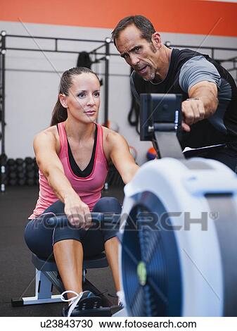Stock Image - Woman exercising on row machine supervised by her trainer. Fotosearch