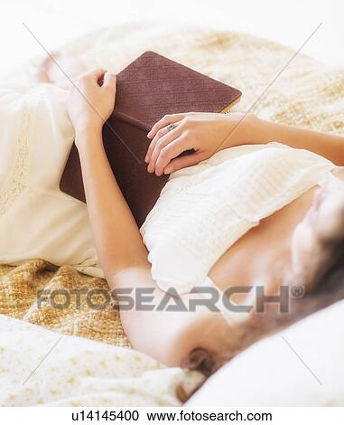 Stock Image - Woman in white chemise laying in bed and holding book. Fotosearch