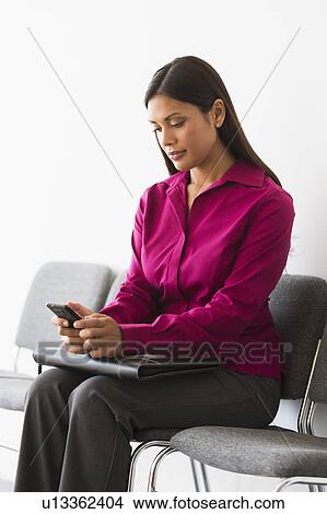 Picture - Woman sitting in waiting room and text messaging. Fotosearch