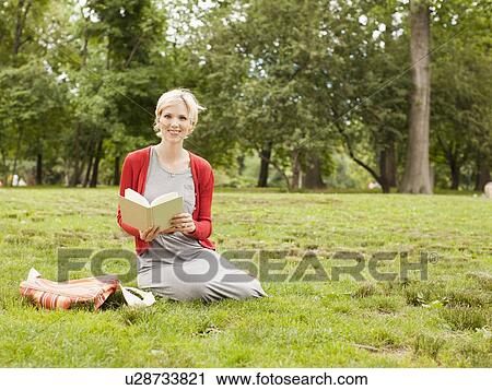 Stock Image - Woman sitting on grass in park holding book. Fotosearch