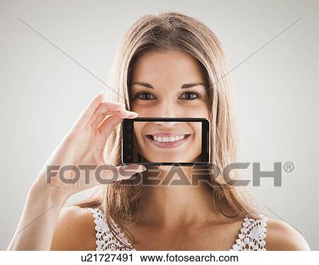 Stock Image - Woman smiling with smart phone in front of mouth, smile showing on screen of smart phone. Fotosearch