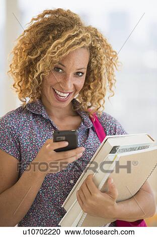 Stock Image - Young woman carrying books and using mobile phone. Fotosearch