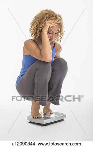 Picture - Young woman crouching on weight scale, studio shot. Fotosearch