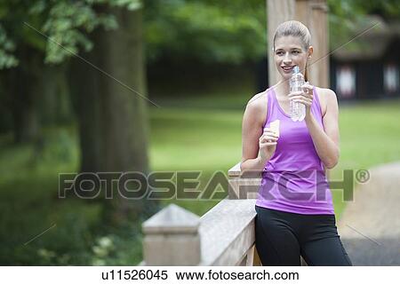 Stock Photography - Young woman leaning against bridge after running and eating cookie and drinking water from bottle.. Fotosearch