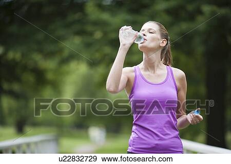 Stock Photo - Young woman walking over bridge and drinking bottle of water after jogging. Fotosearch