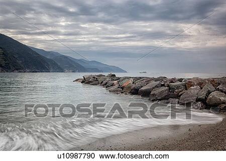 Beach and coastline at Monterosso al Mare: stone breakwater with motion blur of waves, with hills beyond, single boat, early morning with stormy sky, Cinque Terre. Liguria, Italy View Large Photo Image Stock Image - Beach and coastline at Monterosso al Mare: stone breakwater with motion blur of waves, with hills beyond, single boat, early morning with stormy sky, Cinque Terre. Liguria, Italy. Fotosearch