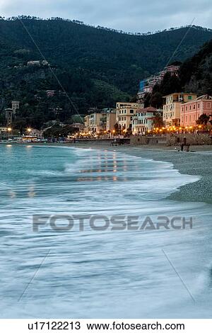 Stock Image - Beach and coastline at Monterosso al Mare: sea front of town with motion blur of waves, with hills beyond, illuminations reflected on beach, Cinque Terre. Liguria, Italy. Fotosearch