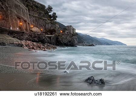 Stock Image - Beach and coastline at Monterosso al Mare: pink rocks at sea edge with motion blur of waves, single house perched above, stormy sunset sky with night illuminations reflected on beach, Cinque Terre. Liguria, Italy. Fotosearch