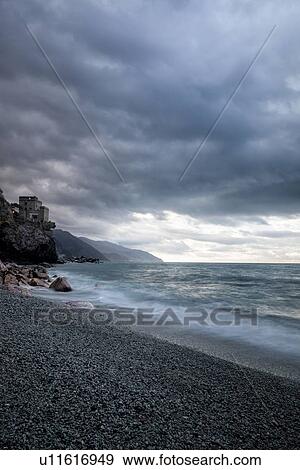 Stock Photo - Beach and coastline at Monterosso al Mare: pink rocks at sea edge with motion blur of waves, single house perched above, and stormy sunset sky, Cinque Terre. Liguria, Italy. Fotosearch