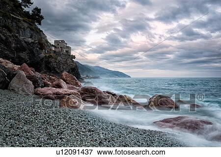 Beach and coastline at Monterosso al Mare: pink rocks at sea edge with motion blur of waves, single house perched above, and stormy sunset sky, Cinque Terre. Liguria, Italy View Large Photo Image Stock Photo - Beach and coastline at Monterosso al Mare: pink rocks at sea edge with motion blur of waves, single house perched above, and stormy sunset sky, Cinque Terre. Liguria, Italy. Fotosearch