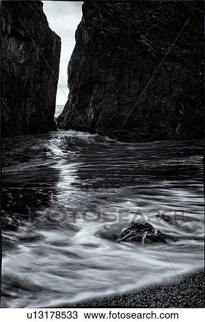 Stock Image - Beach at Monterosso al Mare: imposing black rock close up, at sea edge with motion blur of waves, grey sky, small rocks in foreground, monochrome, Cinque Terre. Liguria, Italy. Fotosearch