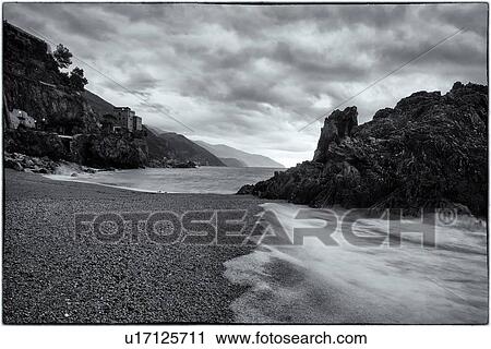 Stock Image - Beach at Monterosso al Mare: imposing black rock at sea edge with motion blur of waves, grey sky, coastline with hills beyond, monochrome image, Cinque Terre. Liguria, Italy. Fotosearch