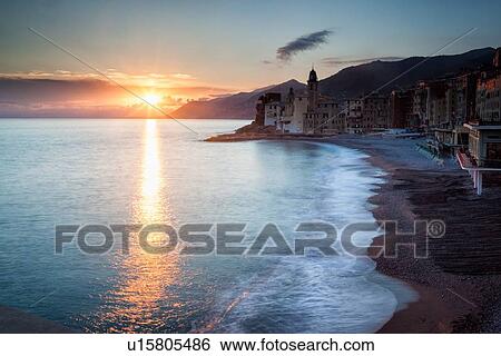 Stock Photograph - Camogli: beach and sea with rolling waves, church tower and sea front buildings, sunset sky with dramatic clouds and sunstar, Liguria, Italy. Fotosearch