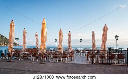 Stock Image - Camogli in springtime: empty terrace cafe with closed umbrellas in soft pink sunset light, iron lamps and railing, calm sea beyond, Liguria, Italy. Fotosearch