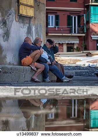 Stock Photo - Camogli: three men sitting on stone wall chatting with old cracked stucco wall and red houses, street beyond, yellow wooden window shutter above, all reflected in large puddle, Liguria, Italy. Fotosearch