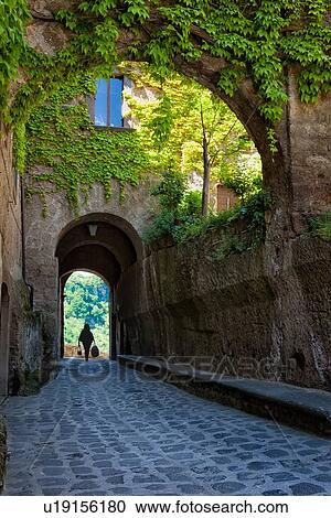 Cività di Bagnoregio: centre of village, with ivy covered stone houses, single woman silhouetted as she walks through cobblestone arched walkway leading to bridge and valley with trees beyond, Umbria, Italy View Large Photo Image Stock Image - Cività di Bagnoregio: centre of village, with ivy covered stone houses, single woman silhouetted as she walks through cobblestone arched walkway leading to bridge and valley with trees beyond, Umbria, Italy. Fotosearch