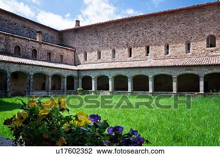 Stock Image - Cloister of San Fortunato, Todi with view of arched stone portico with red tiled roof, and red brick cells above, blue sky in springtime, pansies in foreground, Umbria, Italy. Fotosearch
