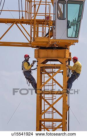 Picture - Close up of horizontal part of yellow crane with two men attached below the cabin by chains, wearing yellow jackets, and red safety helmets, hammering as they work on crane assembly, Umbria, Italy. Fotosearch