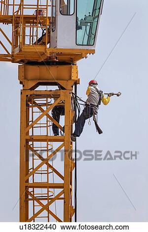 Close up of horizontal part of yellow crane with two men attached below the cabin by chains, wearing yellow jackets, and red safety helmets, hammering as they work on crane assembly, Umbria, Italy View Large Photo Image Stock Image - Close up of horizontal part of yellow crane with two men attached below the cabin by chains, wearing yellow jackets, and red safety helmets, hammering as they work on crane assembly, Umbria, Italy. Fotosearch